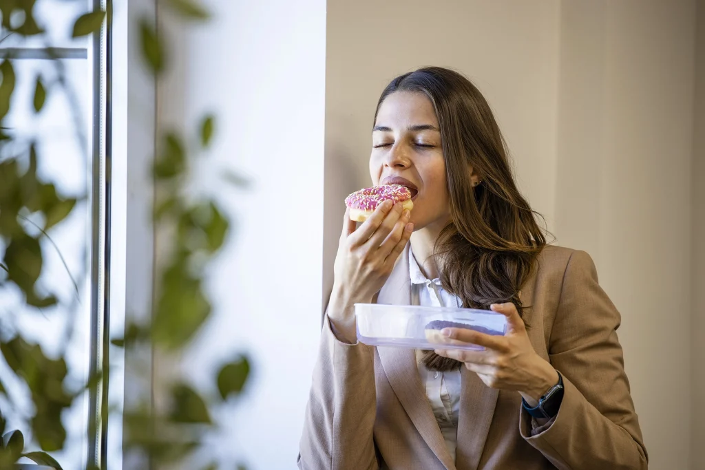 Femme mangeant un beigne au travail, illustrant le fait de manger ses émotions et la recherche de réconfort par la nourriture.