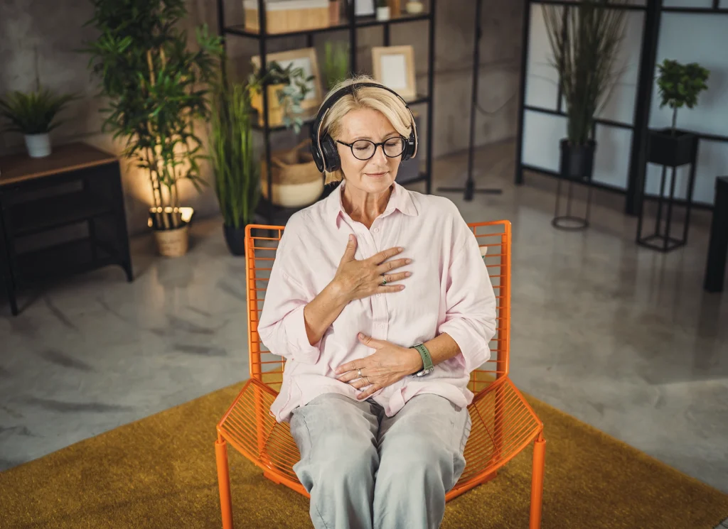 Femme assise, les yeux fermés, une main sur la poitrine et l’autre sur le ventre, pratiquant un exercice de respiration consciente avec des écouteurs.